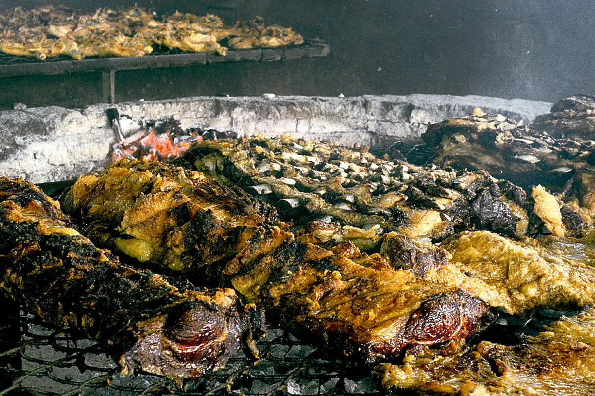 Beef ribs and other cuts of meat roasting on an iron grill beside a bed of embers in a brick-lined asado pit